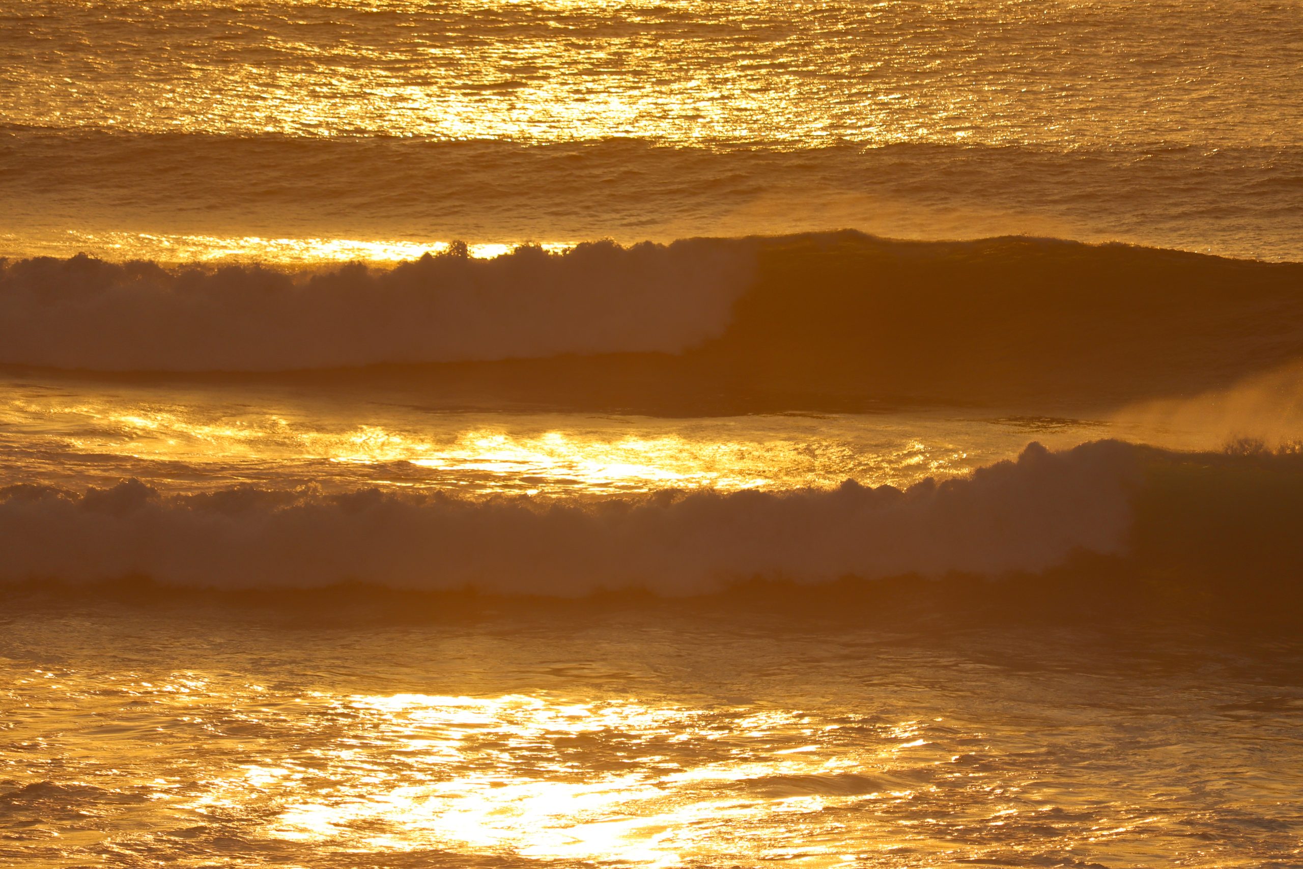 Alternative Text: Goldene Wellen brechen im warmen Abendlicht der untergehenden Sonne vor der Küste von Bali.
