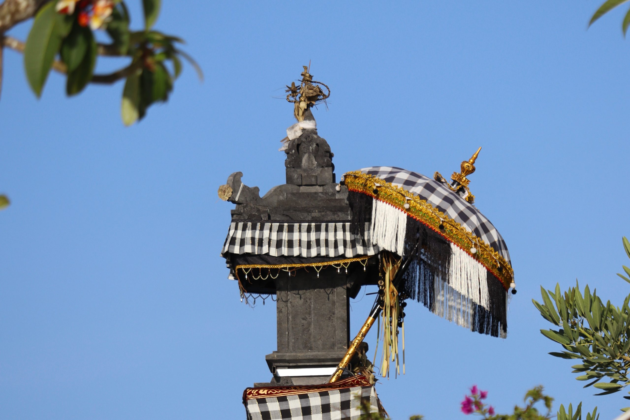 Traditioneller balinesischer Hausschrein (Penunggun Karang) mit schwarz-weiss kariertem Poleng-Tuch und Schirm vor blauem Himmel auf Bali.