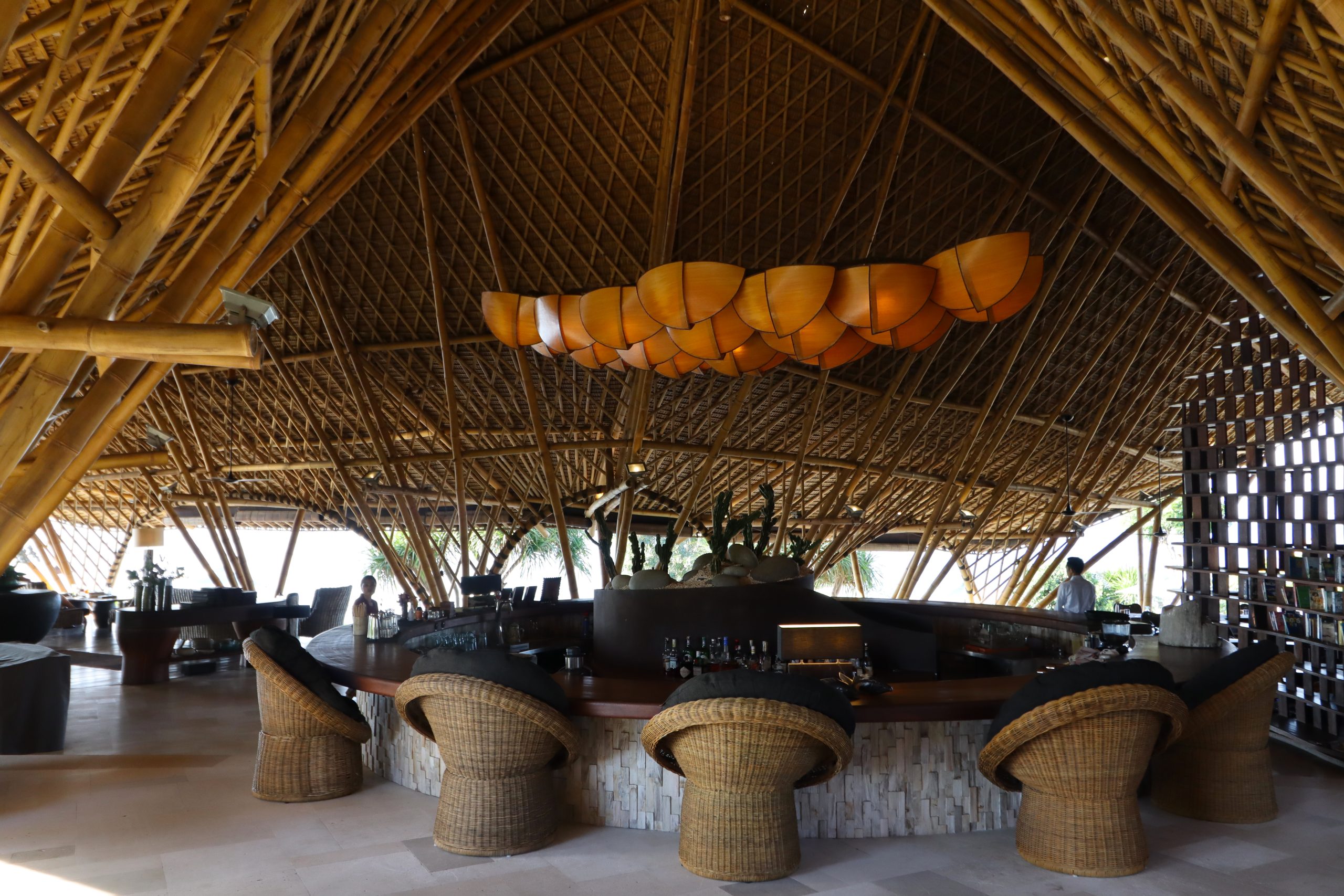 Alternative Text: Nachhaltige Bambusarchitektur und Bar-Lounge im Suarga Padang Padang Resort auf Bali mit gewölbter Dachkonstruktion und geflochtenen Sesseln.