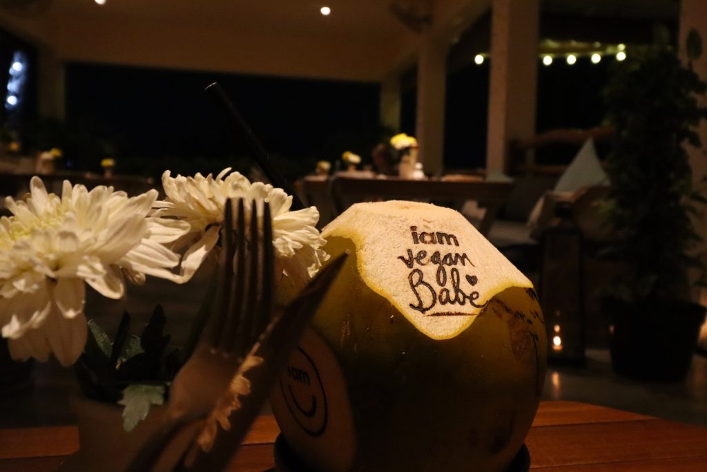 Alternative Text: Frische Trinkkokosnuss mit eingraviertem Schriftzug I am Vegan Babe und weissen Blumen in einem Restaurant in Canggu auf Bali.