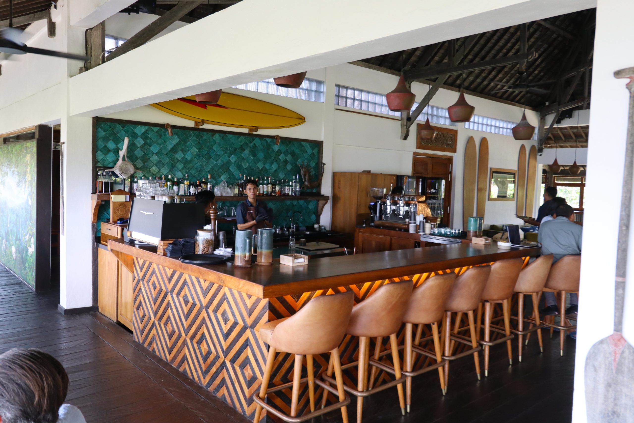 Alternative Text: Barbereich des Uluwatu Surf Villas Restaurants auf Bali mit Barhockern und einem Surfboard mit Blitz-Logo an der Wand.